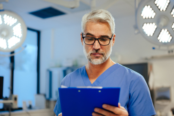 Portrait of handsome male doctor in scrubs. ER doctor examining patient, reading medical test, lab results in clipboard. ER with patient with life threatening health problems.