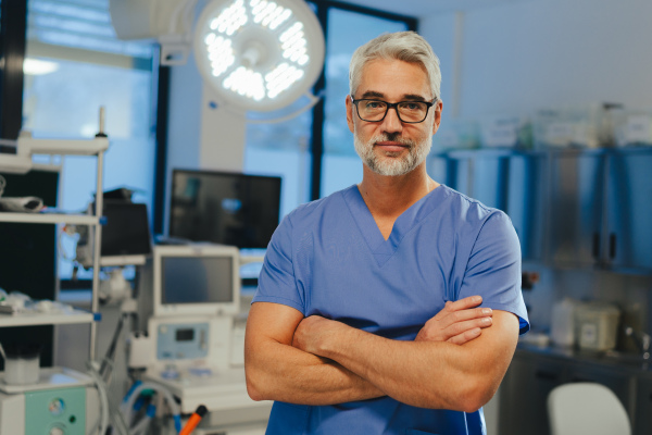 Portrait of confident ER doctor standing in hospital, emergency room. Handsome doctor in scrubs holding clipboard with test results, standing in modern private clinic, looking at camera.