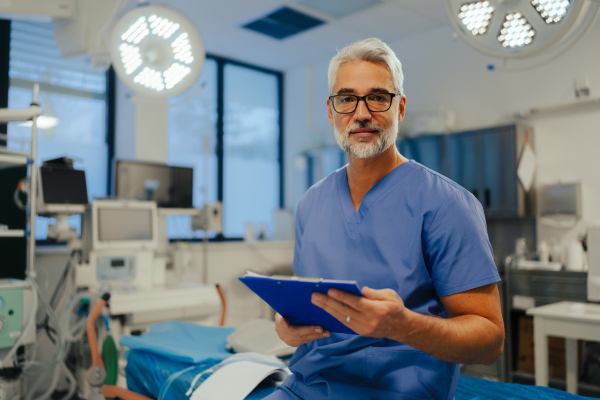 Portrait of confident ER doctor standing in hospital, emergency room. Handsome doctor in scrubs holding clipboard with test results, standing in modern private clinic, looking at camera.