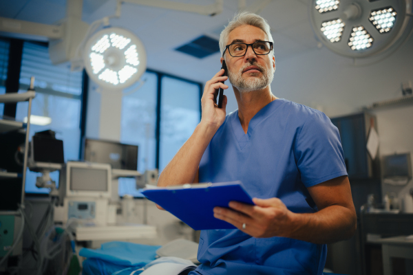 Portrait of confident ER doctor standing in hospital, emergency room and phone calling. Handsome doctor in scrubs holding clipboard with test results, standing in modern private clinic.