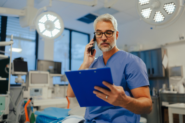 Portrait of confident ER doctor standing in hospital, emergency room. Handsome doctor in scrubs holding clipboard with test results, standing in modern private clinic, looking at camera.