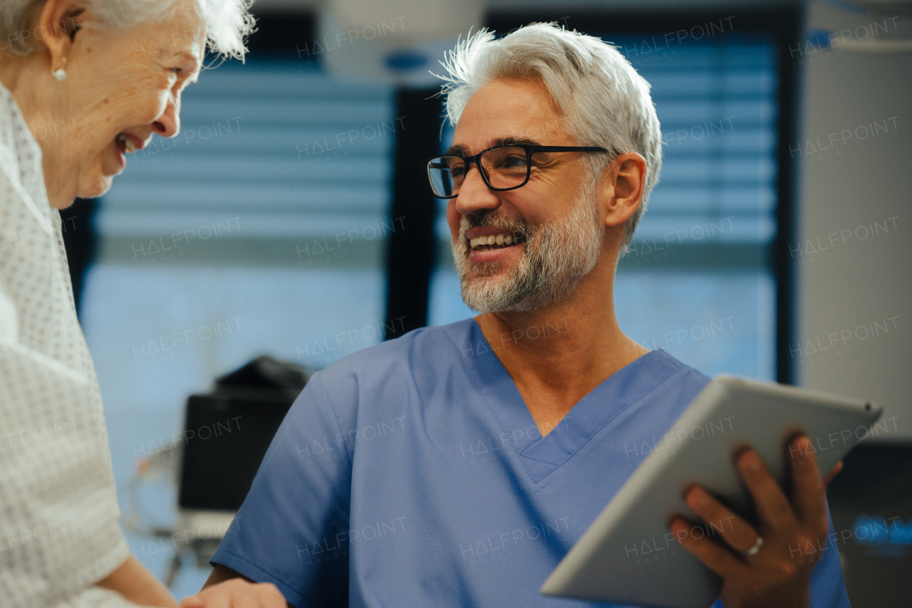 Portrait of handsome mature doctor talking with elderly patient in hospital. Consulting medical test, lab results, smiling, laughing. Emotional support for elderly woman.