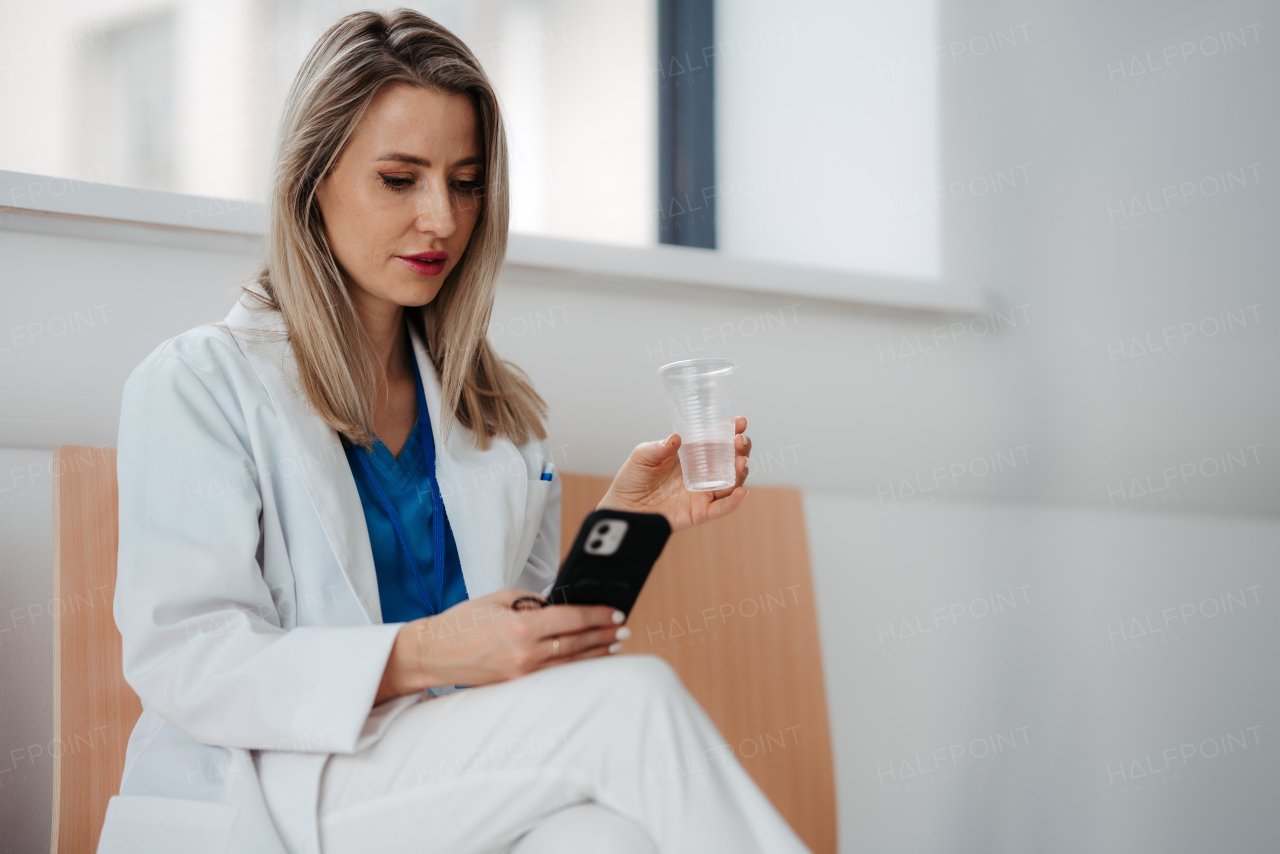 Beautiful female doctor is taking a break during her work shift at hospital, drinking water and scrolling on smartphone.