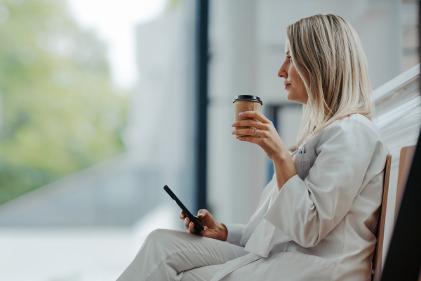 Beautiful female doctor is taking a break during her work shift at hospital, drinking coffee and scrolling on smartphone. Side view.