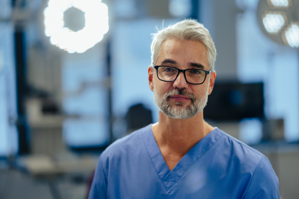 Portrait of confident ER doctor standing in hospital emergency room. Handsome doctor in blue scrubs standing in modern private clinic, looking at camera.