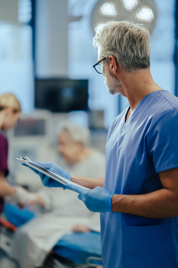 Mature male doctor looking at patient in hospital bed. ER doctor examining senior patient, reading her medical test, lab results in clipboard. ER with patient with life threatening health problems.