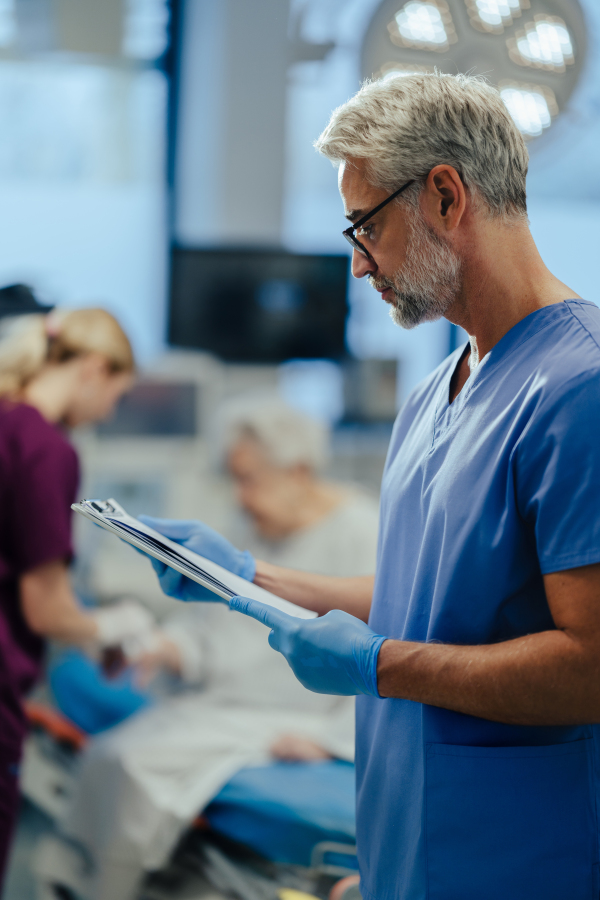 Portrait of handsome male doctor, patient in hospital bed behind. ER doctor examining senior patient, reading her medical test, lab results in clipboard.ER with patient with life threatening health problems.