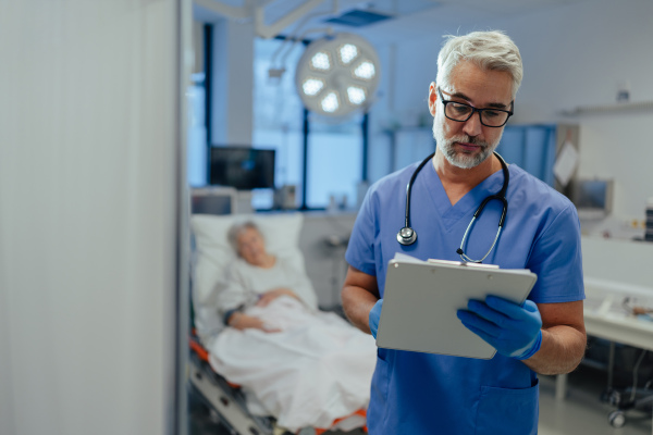 Portrait of confident ER doctor standing in hospital, emergency room. Handsome doctor in scrubs holding clipboard with test results, standing in modern private clinic, looking at camera.