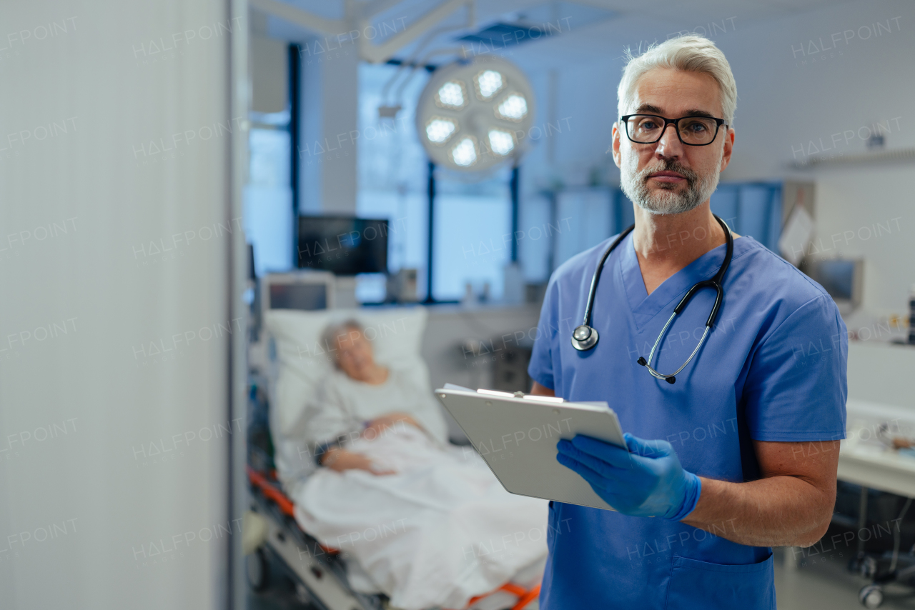 Portrait of handsome male doctor, patient in hospital bed behind. ER doctor examining senior patient, reading her medical test, lab results in clipboard.ER with patient with life threatening health problems.