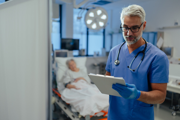 Portrait of handsome male doctor, patient in hospital bed behind. ER doctor examining senior patient, reading her medical test, lab results in clipboard.ER with patient with life threatening health problems.