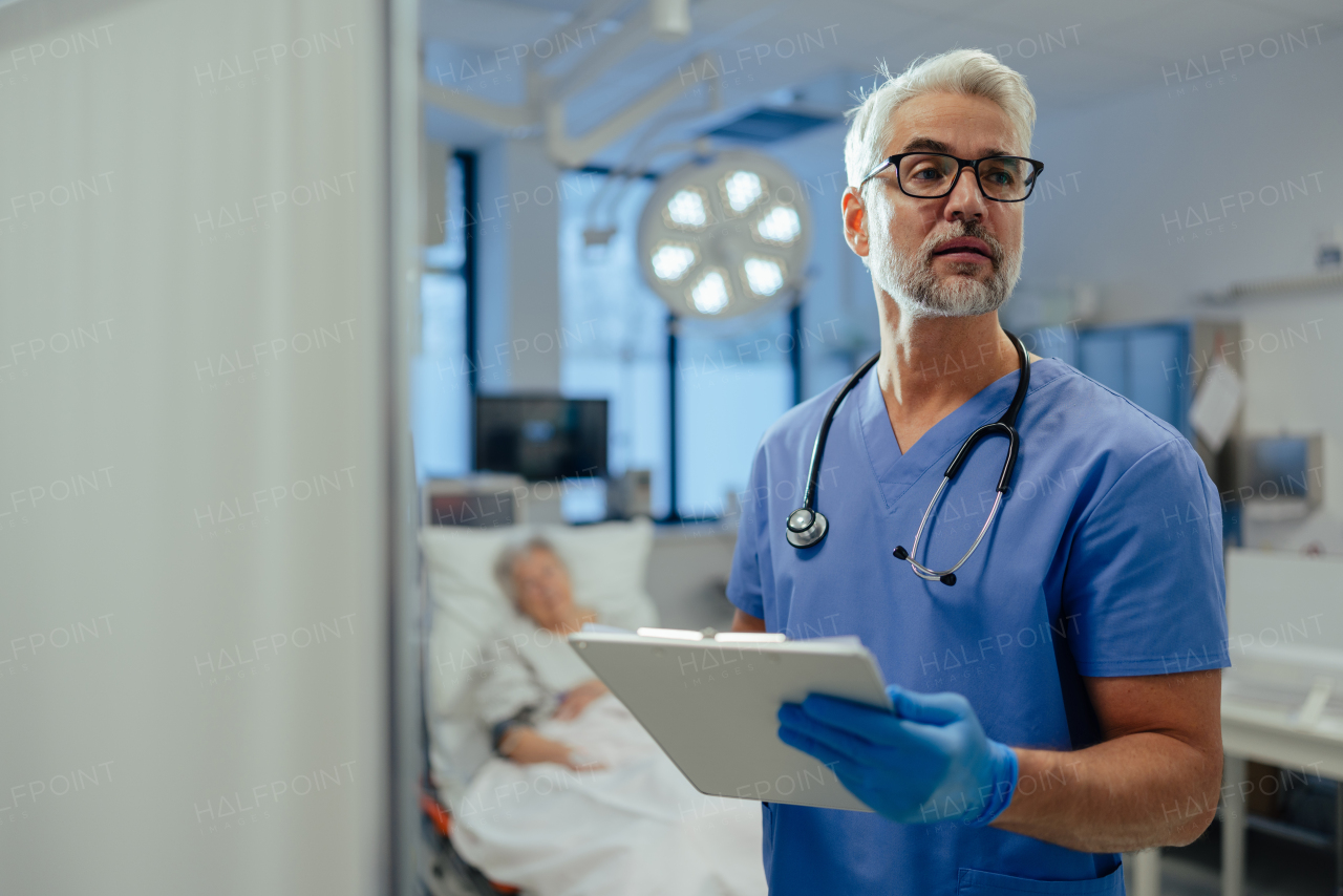 Portrait of handsome male doctor, patient in hospital bed behind. ER doctor examining senior patient, reading her medical test, lab results in clipboard.ER with patient with life threatening health problems.