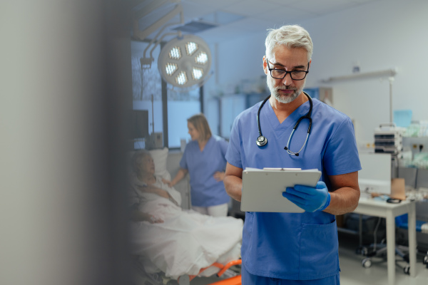 Portrait of handsome male doctor, patient in hospital bed behind. ER doctor examining senior patient, reading her medical test, lab results in clipboard.ER with patient with life threatening health problems.