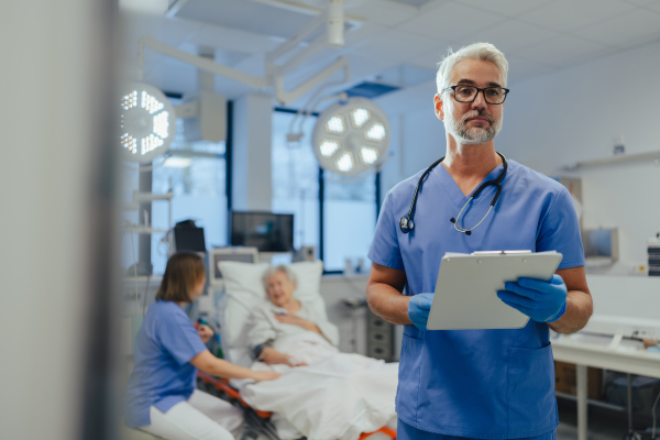 Portrait of handsome male doctor, patient in hospital bed behind. ER doctor examining senior patient, reading her medical test, lab results in clipboard.ER with patient with life threatening health problems.