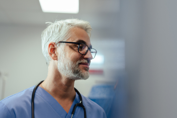 Portrait of confident mature doctor in hospital room. Handsome doctor with gray hair wearing medical scrubs, eyeglasses and stethoscope around neck standing in modern private clinic, talking with patient, nurse.