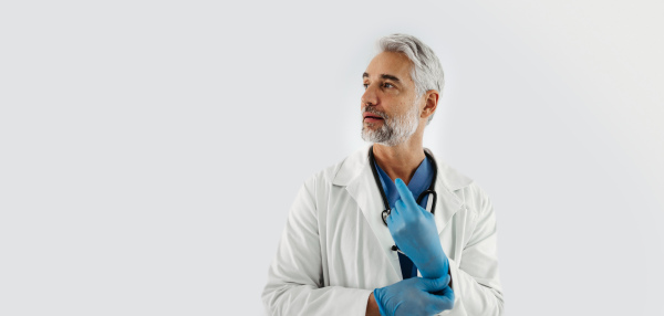 Confident mature doctor putting on medical or examination gloves. Handsome doctor with gray hair wearing white coat, stethoscope around neck. Banner, white background with copy space.