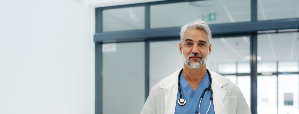 Portrait of confident mature doctor standing in Hospital corridor. Handsome doctor with gray hair wearing white coat, scrubs, stethoscope around neck standing in modern private clinic. Medical banner.