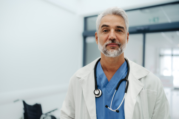 Portrait of confident mature doctor standing in Hospital corridor. Handsome doctor with gray hair wearing white coat, scrubs, stethoscope around neck standing in modern private clinic, looking at camera.