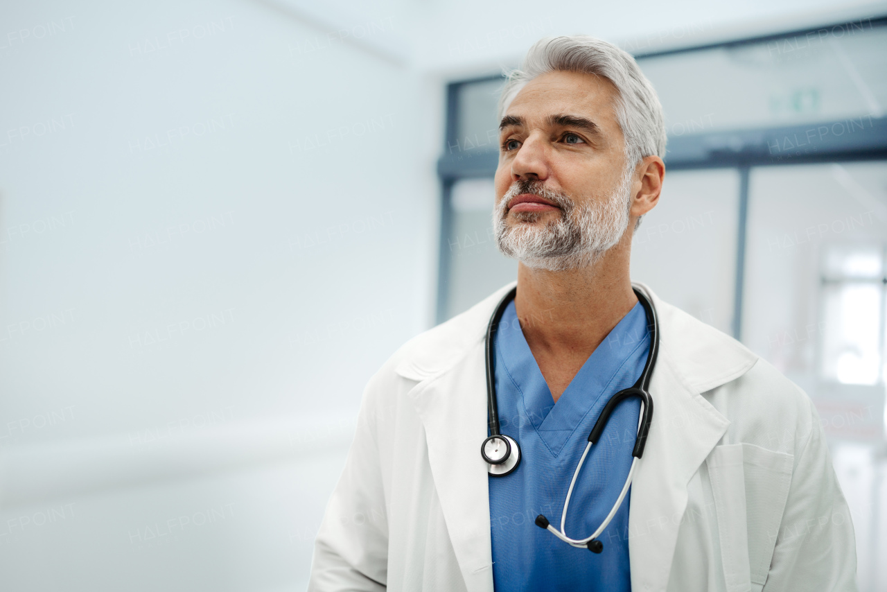 Portrait of confident mature doctor standing in Hospital corridor. Handsome doctor with gray hair wearing white coat, scrubs, stethoscope around neck standing in modern private clinic, looking at camera.