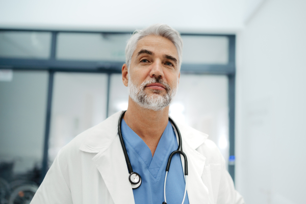 Portrait of confident mature doctor standing in Hospital corridor. Handsome doctor with gray hair wearing white coat, scrubs, stethoscope around neck standing in modern private clinic, looking at camera.