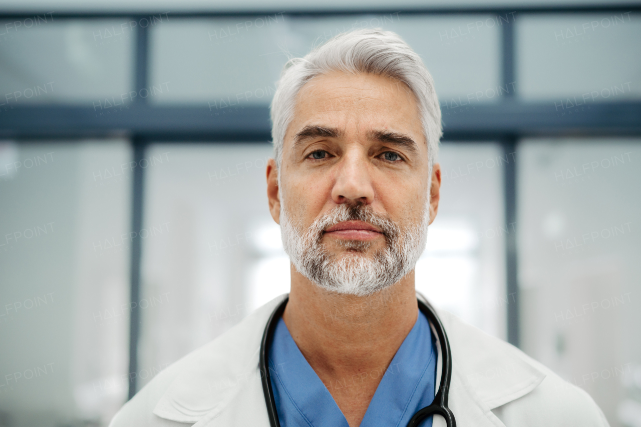 Portrait of confident mature doctor standing in Hospital corridor. Handsome doctor with gray hair wearing white coat, scrubs, stethoscope around neck standing in modern private clinic, looking at camera.