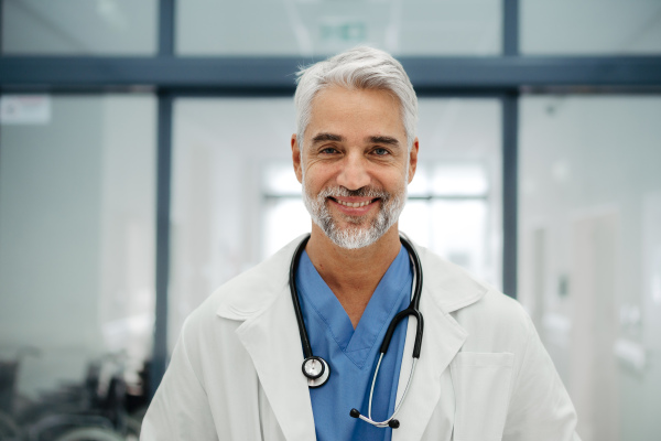 Portrait of confident mature doctor standing in Hospital corridor. Handsome doctor with gray hair wearing white coat, scrubs, stethoscope around neck standing in modern private clinic, looking at camera.