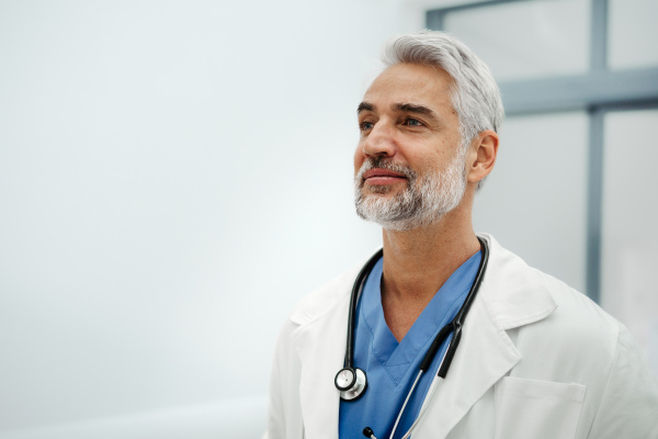 Portrait of confident mature doctor standing in Hospital corridor. Handsome doctor with gray hair wearing white coat, scrubs, stethoscope around neck standing in modern private clinic, looking at camera.