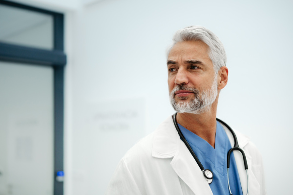 Portrait of confident mature doctor standing in Hospital corridor. Handsome doctor with gray hair wearing white coat, scrubs, stethoscope around neck standing in modern private clinic, looking at camera.