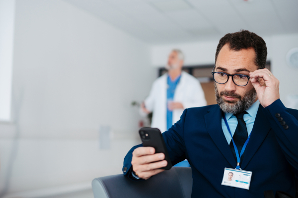 Pharmaceutical sales representative sitting in medical building, waiting for the doctor, presenting new pharmaceutical product. Male drug rep sitting in hall scrolling on smartphone.