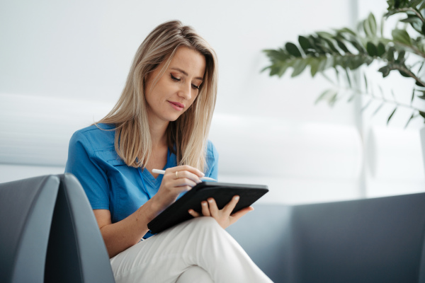 Portrait of female doctor sitting in office, looking at test results, CT scans in tablet. Female surgeon in modern medical clinic using digital technology.