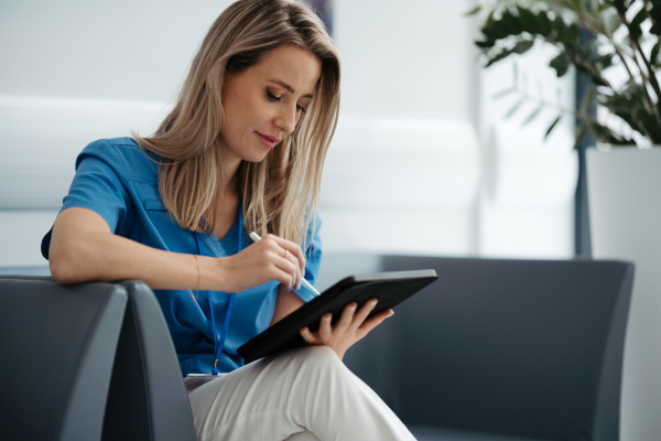 Portrait of female doctor sitting in office, looking at test results, CT scans in tablet. Female surgeon in modern medical clinic using digital technology.