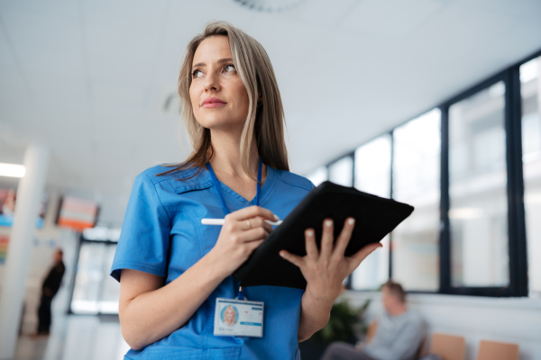 Portrait of confident female doctor standing in Hospital corridor. Beautiful nurse wearing blue scrubs, holding clipboard standing in modern private clinic, low angle photography.