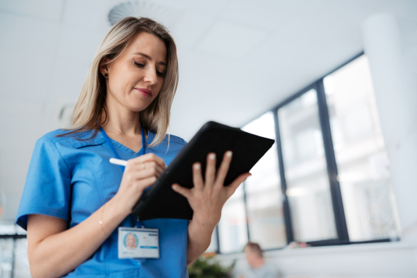 Portrait of confident female doctor standing in Hospital corridor. Beautiful nurse wearing blue scrubs, holding clipboard standing in modern private clinic, low angle photography.