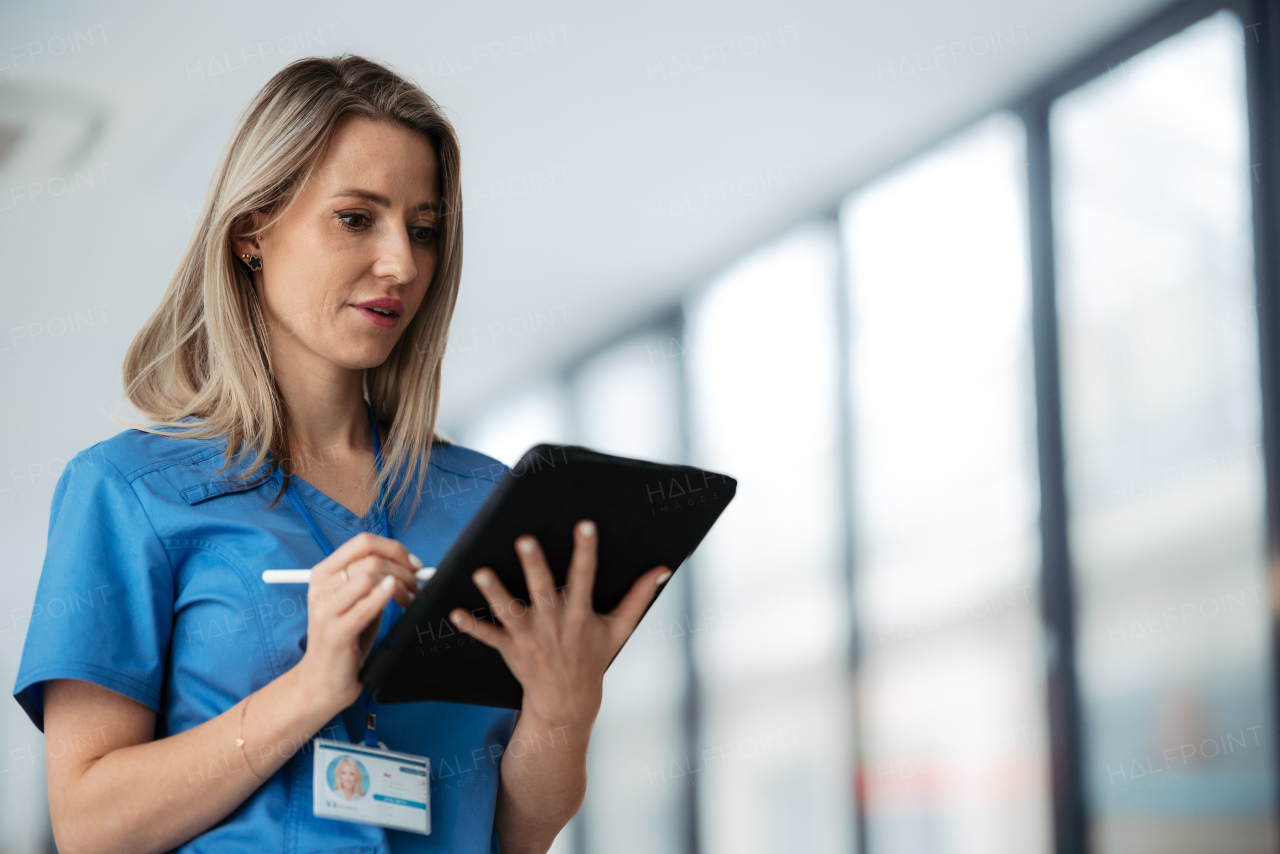 Portrait of confident female doctor standing in Hospital corridor. Beautiful nurse wearing blue scrubs, holding clipboard standing in modern private clinic, low angle photography.