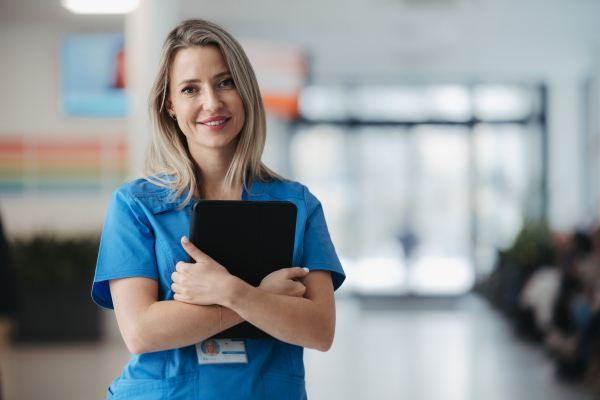 Portrait of confident female doctor in hospital corridor. Beautiful nurse wearing blue uniform, holding clipboard standing in modern private clinic, looking at camera, smiling.