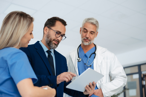 Pharmaceutical sales representative talking with doctors in medical building, presenting them new product on tablet, business talk. Hospital director consulting with healthcare staff in hall.