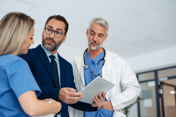 Pharmaceutical sales representative talking with doctors in medical building, presenting them new product on tablet, business talk. Hospital director consulting with healthcare staff in hall.