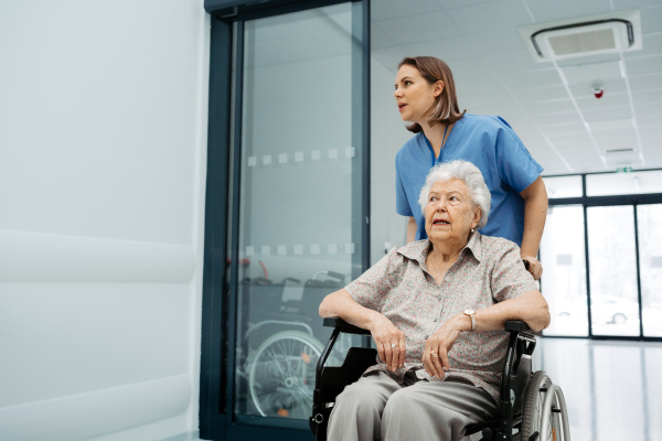 Portrait of nurse pushing senior patient in a wheelchair across hospital corridor, hall. Emotional support for elderly woman.