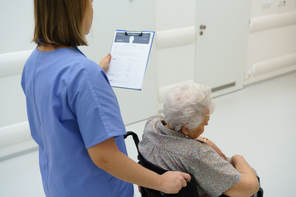 Nurse pushing senior patient in a wheelchair, reading medical document in clipboard, patient records. Emotional support for elderly woman.