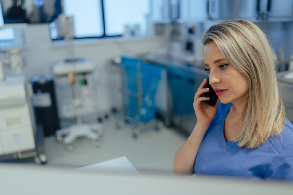 Portrait of ER doctor in hospital phone calling. Remote consultations with patient, telemedicine. Beautiful nurse in scrubs making call and typing on medical computer in emergency room.