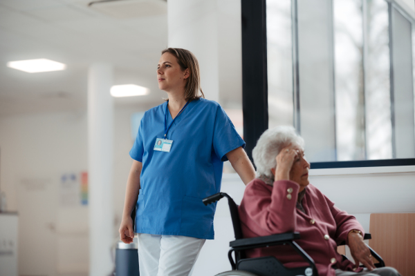 Tired nurse waiting with senior patient in wheelchair in hospital corridor. Emotional support for elderly woman.