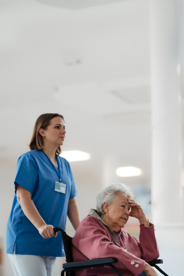 Nurse pushing senior patient in a wheelchair across hospital corridor, hall. Emotional support for elderly woman.