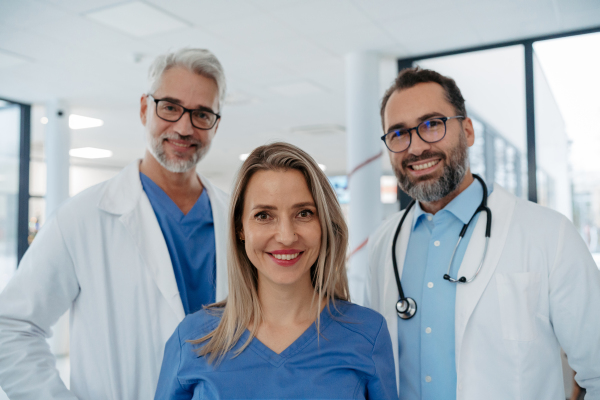 Portrait of confident three doctors standing in Hospital corridor. Medical team wearing white coat, stethoscope around neck standing in modern private clinic, looking at camera, smiling,