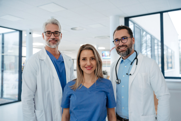 Portrait of confident three doctors standing in Hospital corridor. Medical team wearing white coat, stethoscope around neck standing in modern private clinic, looking at camera, smiling,