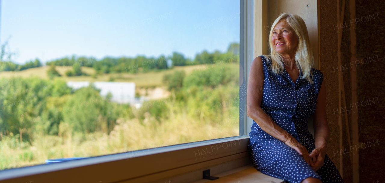 Happy senior woman resting at window, enjoying view inside of her unfinished ecological wooden house.