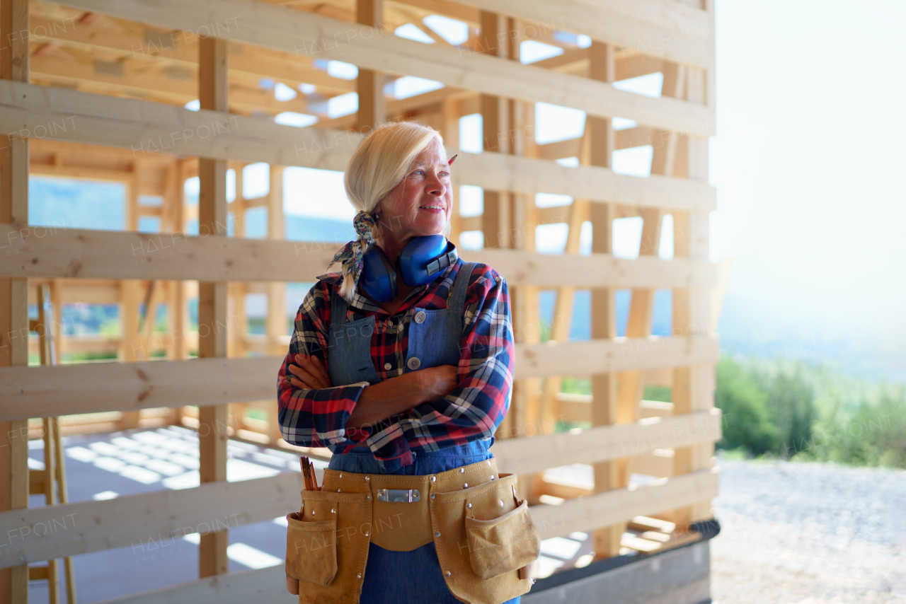 Senior woman in working clothes posing outdoor of her unfinished ecological sustainable wooden house. Concept of active independent women and seniors, eco house and healthy lifestyle.