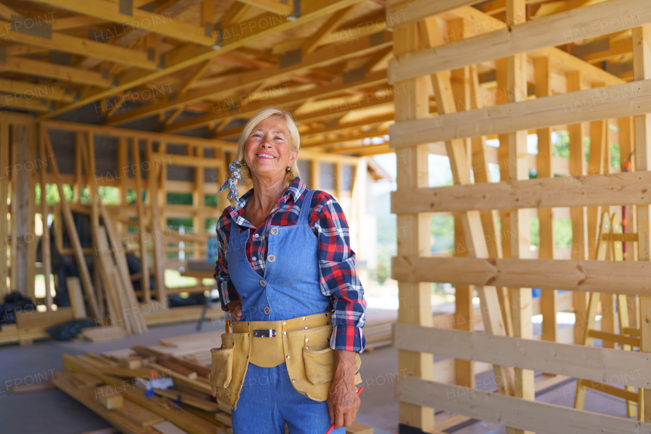 Senior woman in working clothes posing inside her unfinished ecological sustainable wooden house. Concept of active independent women and seniors, eco house and healthy lifestyle.