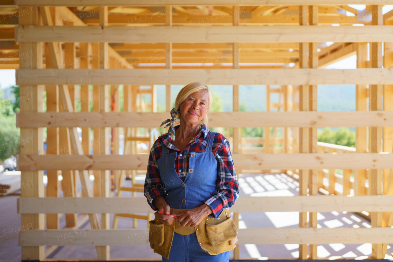 Senior woman in working clothes posing inside her unfinished ecological sustainable wooden house. Concept of active independent women and seniors, eco house and healthy lifestyle.