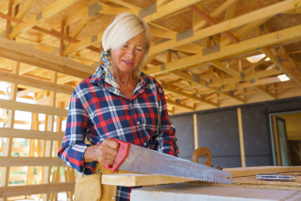 Senior woman sawing inside of her unfinished ecological sustainable wooden eco house. Concept of active independent women and seniors, eco house and healthy lifestyle.