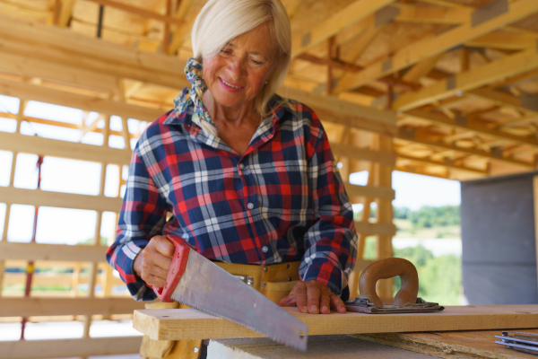 Senior woman sawing inside of her unfinished ecological sustainable wooden eco house. Concept of active independent women and seniors, eco house and healthy lifestyle.