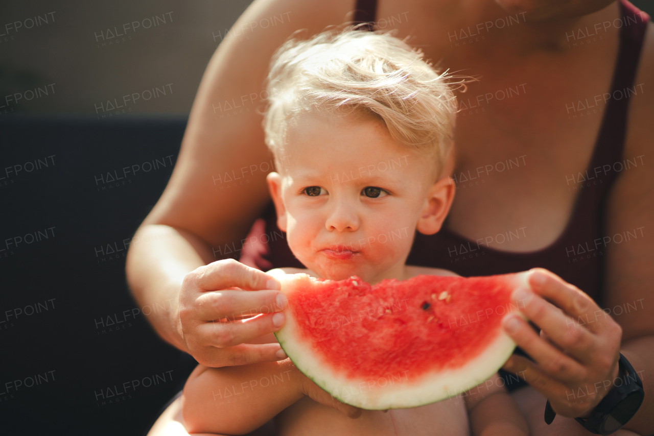 A cute little boy with unrecognizable mother eating watermelon in garden in summer.